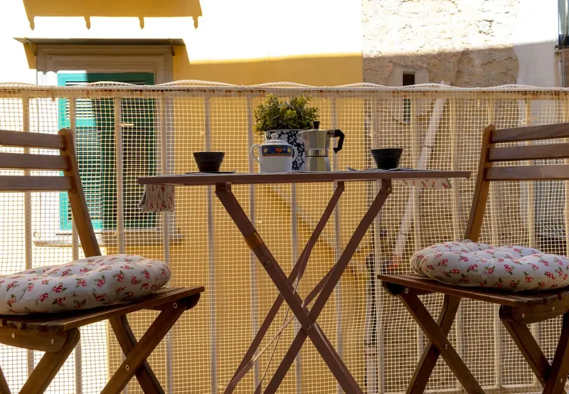 The balcony: two wooden chairs with floral cushions, the little table with the moka pot and cups and, opposite, the yellow façade with green shutters.