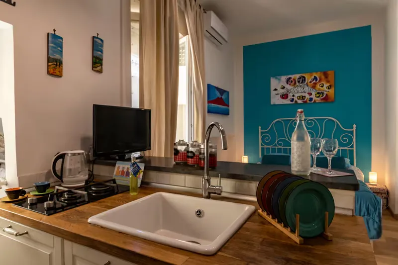The kitchen: the sink, the gas hob and colourful plates on the breakfast bar; behind, the double bed against the turquoise wall.
