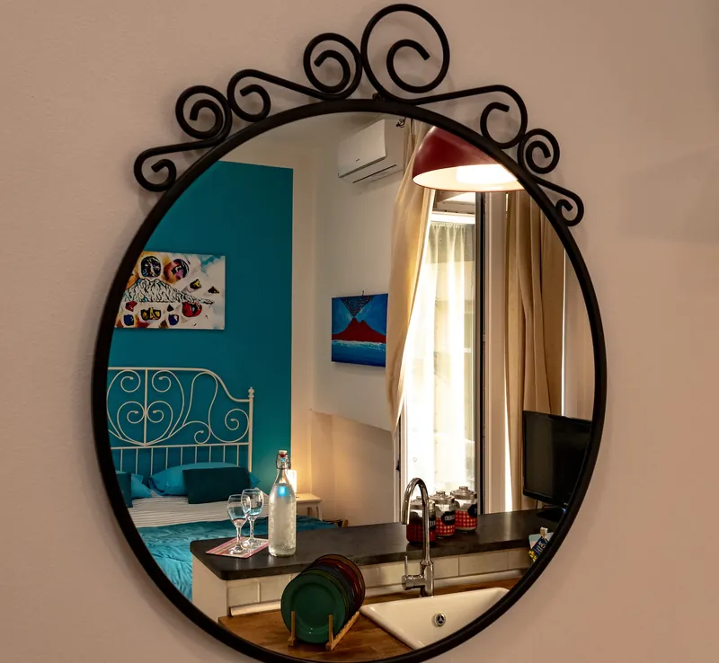 The round wrought-iron mirror, reflecting the bed against the turquoise wall, the red lamp and the curtained window.